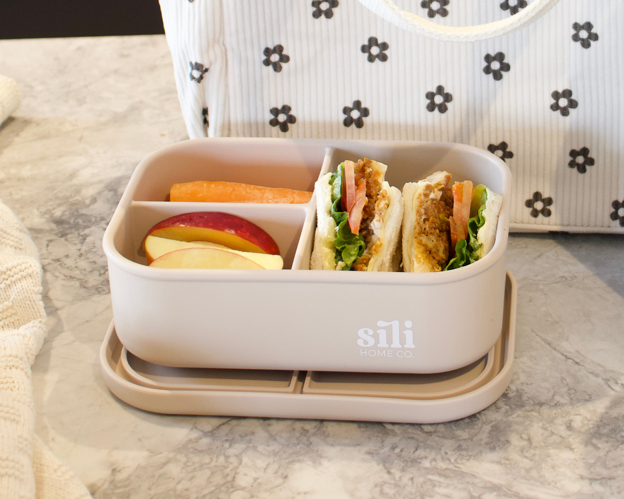 Bento box with food compartments on a marble surface with a patterned bag in the background.